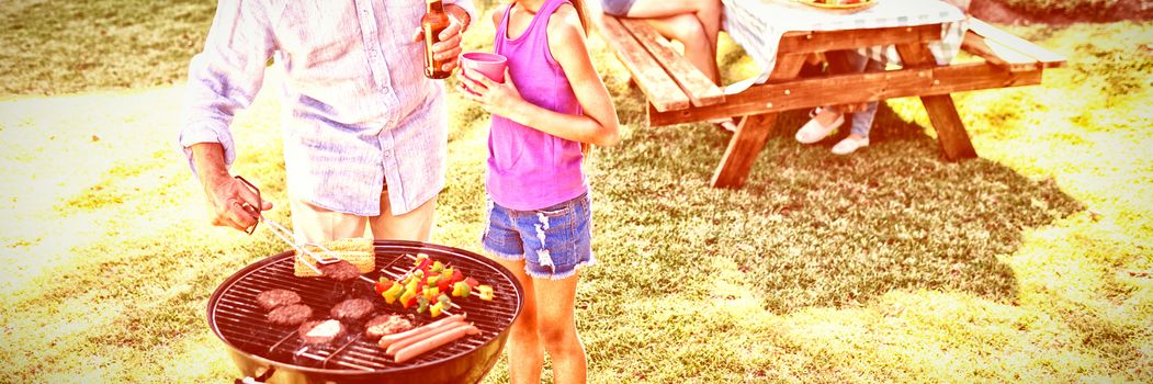 Grandfather and granddaughter preparing barbecue while family having meal in background