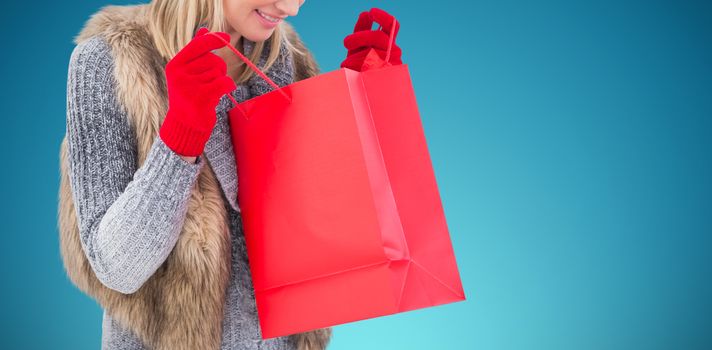 Blonde in winter clothes looking in shopping bag against abstract blue background