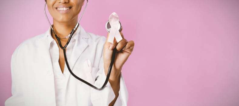 Smiling nurse wearing breast cancer awareness holding pink ribbon on a pink background