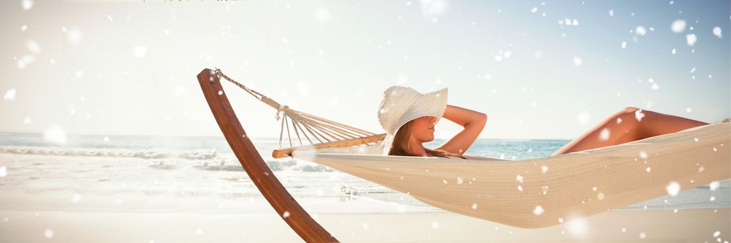Snow falling against woman wearing sunhat and bikini relaxing on hammock