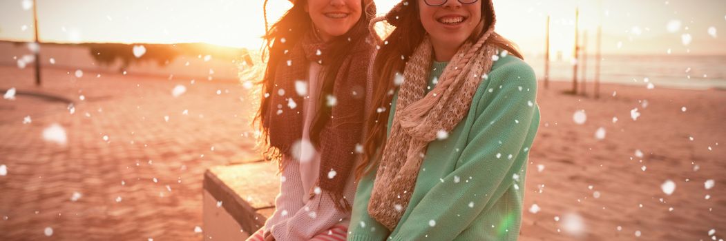 Snow falling against  female friends sitting together on bench at beach Female friends sitting together on bench at beach during sunset