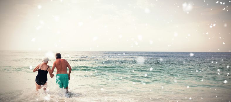 Snow falling against rear view of senior couple holding hands and walking in sea