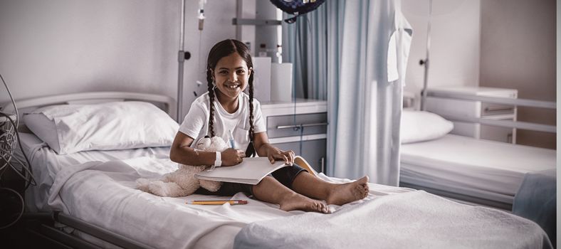 Smiling girl drawing picture in a book while sitting on hospital bed 