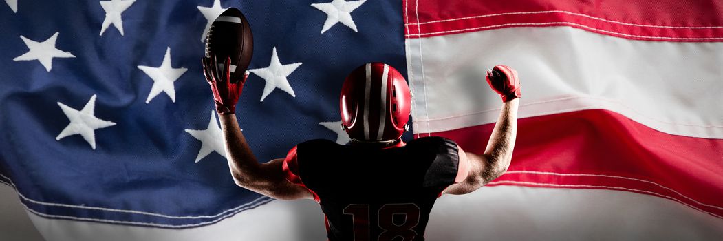 American football player standing with helmet holding football in victory against full frame of american flag