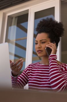Front view of a beautiful  African American woman talking on mobile phone while using her laptop on her terrace at home
