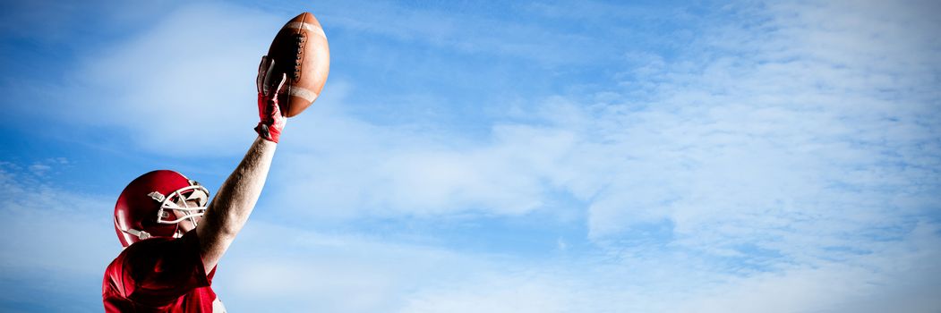 American football player in helmet holding rugby ball with hand raised against low angle view of cloudy sky 