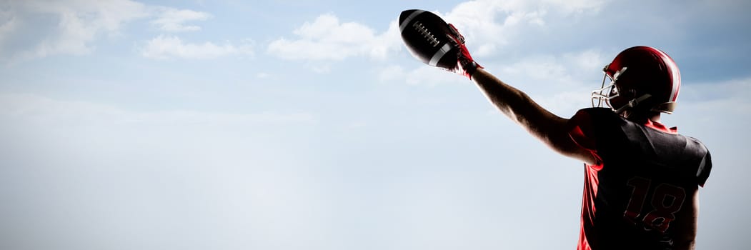 American football player in helmet holding rugby ball with arms in the air against blue sky