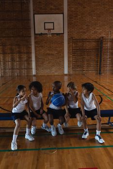 Front view of schoolkids talking with each other and sitting on bench at basketball court in school