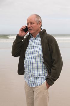 Side view of active senior man talking on mobile phone standing at beach with mountains in the background. He seems happy