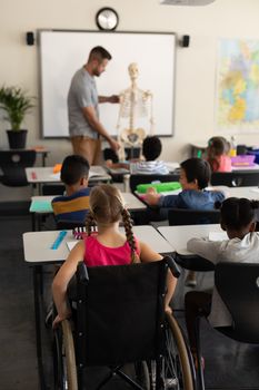 Rear view of disable schoolgirl with classmate studying in classroom sitting at desk of elementary school