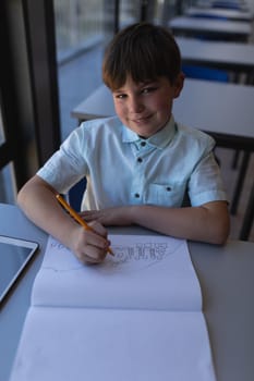 Front view of schoolboy drawing on notebook and looking at camera at desk in classroom of elementary school