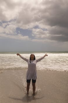 Front view of young woman with arms stretched out standing on beach in the sunshine