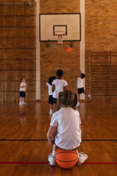 Rear view of schoolgirl sitting on basketball at basketball court in school
