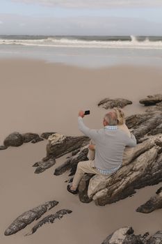 Front view of active Senior couple taking selfie with mobile phone standing on rocks at the beach with ocean in the background. They seem happy