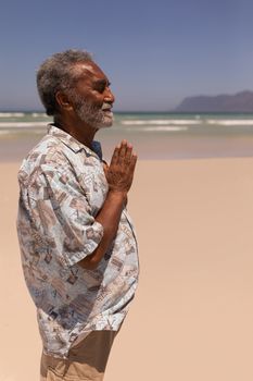 Side view of senior black man with hands clasped praying on beach in the sunshine