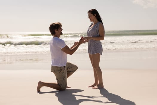 Side view of romantic young man proposing to a woman on his knee at beach in the sunshine