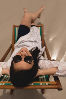 Overhead of beautiful young woman with hand behind head sitting on sun lounger and looking at camera on beach