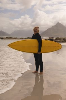 Side view of an active senior woman standing with surfboard on the beach