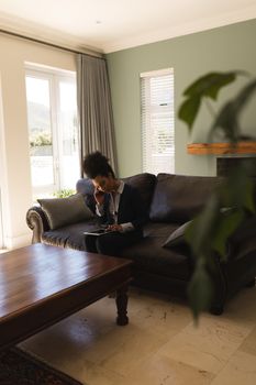 Side view of a female African American real estate agent talking on mobile phone while using digital tablet on a sofa in living room at home