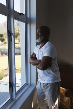 Side view of a thoughtful handsome senior African American man looking through the window witch crossed arms at home