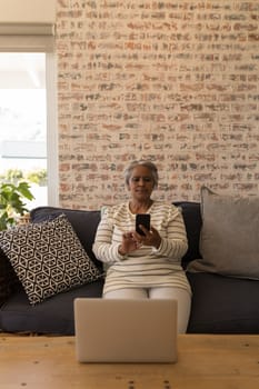 Front view of an active African American senior woman using mobile phone while sitting on a sofa in living room at home