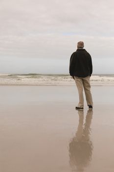 Back view of active senior man standing front of ocean the hands in his pocket. He seems relaxed