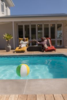 Front view of an active African American senior couple toasting glasses of champagne while relaxing on the lounger chairs in front of the swimming pool in the backyard of home