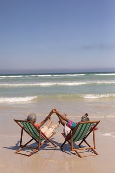Rear view of senior couple relaxing on sun lounger and holding hands on beach in the sunshine