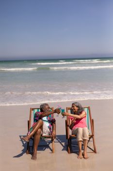High angle view of senior couple relaxing on sun lounger and toasting cocktail glasses on beach in the sunshine