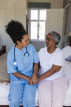 Front view of young pretty African American female doctor comforting senior African American woman in bedroom at home