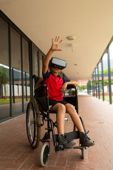 Front view of mixed-race disabled schoolboy using virtual reality headset with one hand in the air in corridor at elementary school