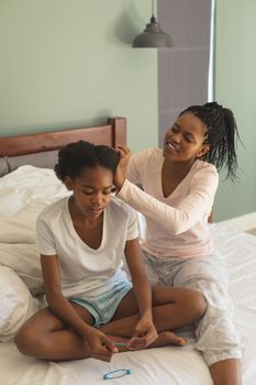 Front view of happy African American mother hairdressing daughter on bed in bedroom at home