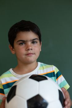 Close-up of a Caucasian schoolboy with a football standing in front of the blackboard in classroom at elementary school