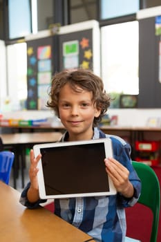 Front view of a Caucasian schoolboy holding digital tablet and looking at the camera in the library at school