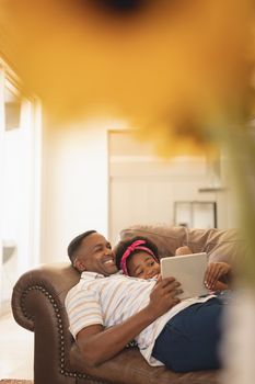 Side view of happy African American father and daughter relaxing on the sofa and using digital tablet at home