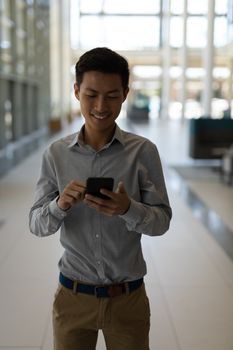 Front view of smiling young Asian male executive using mobile phone in modern office