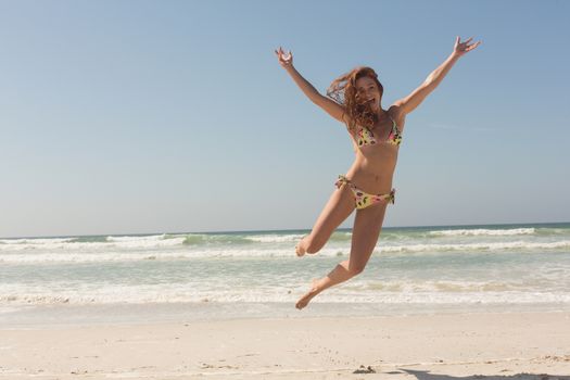 Portrait of happy beautiful young Caucasian woman in bikini jumping on the beach. She is smiling and looking at camera