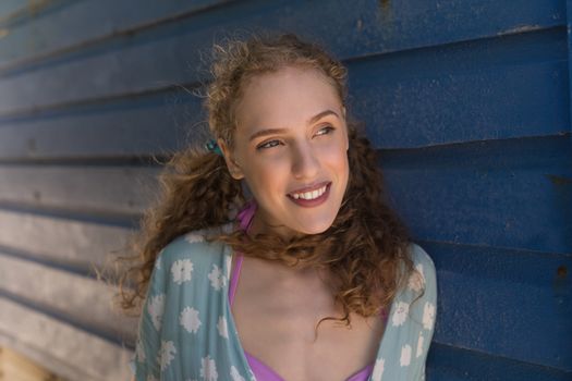 Front view of gorgeous young Caucasian woman smiling and leaning standing against beach hut at beach. she is smiling and looking away