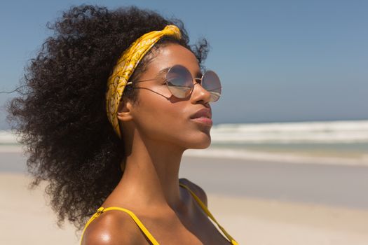Side view of pretty young African American woman in yellow bikini and sunglasses standing on the beach in the sunshine. She is smiling