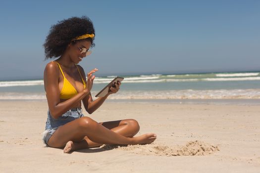 Side view of young African American woman in yellow bikini and sunglasses using digital tablet sitting on the beach. She is smiling
