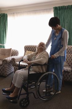 Front view of female doctor interacting with disabled senior woman on wheelchair in living room at home