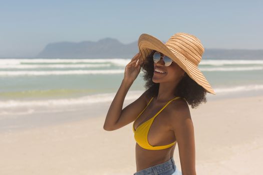 Front view of happy young African American woman in yellow bikini, hat and sunglasses standing on the beach in the sunshine. She is smiling