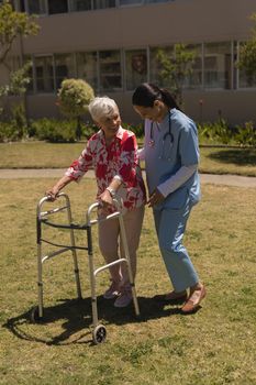 Front view of young happy female doctor helping disabled senior woman in garden on sunny day