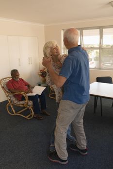 Side view of active senior couple dancing with a senior man reading on a chair at nursing home