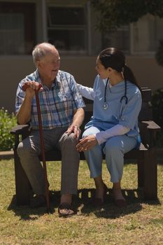 Front view of young female doctor talking with senior man with his cane  while sitting on bench in garden