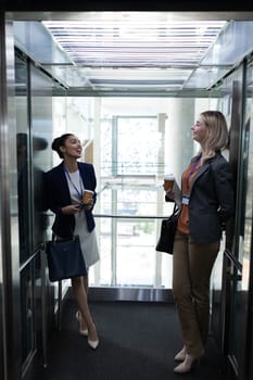 Front view of happy young multi-ethnic businesswomen coffee cup interacting with each other in modern office elevator. They are smiling