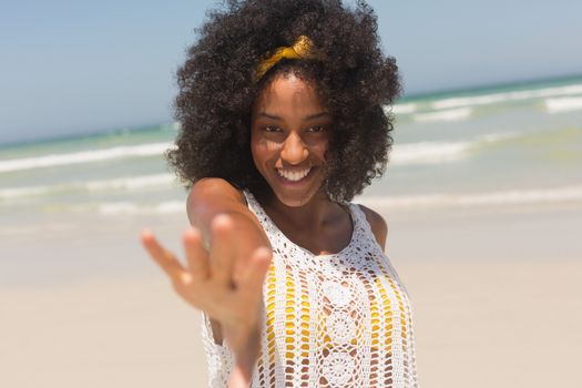 Portrait of happy young African American woman hand reaching towards the camera standing on the beach. She is smiling