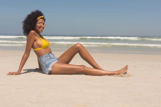Side view of happy young African American woman in yellow bikini and sunglasses relaxing on the beach