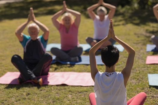 Rear view of female trainer training senior people in performing yoga on yoga mat at park