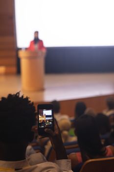Rear view of African-American man with smartphone taking video clip of businesswoman in the auditorium 